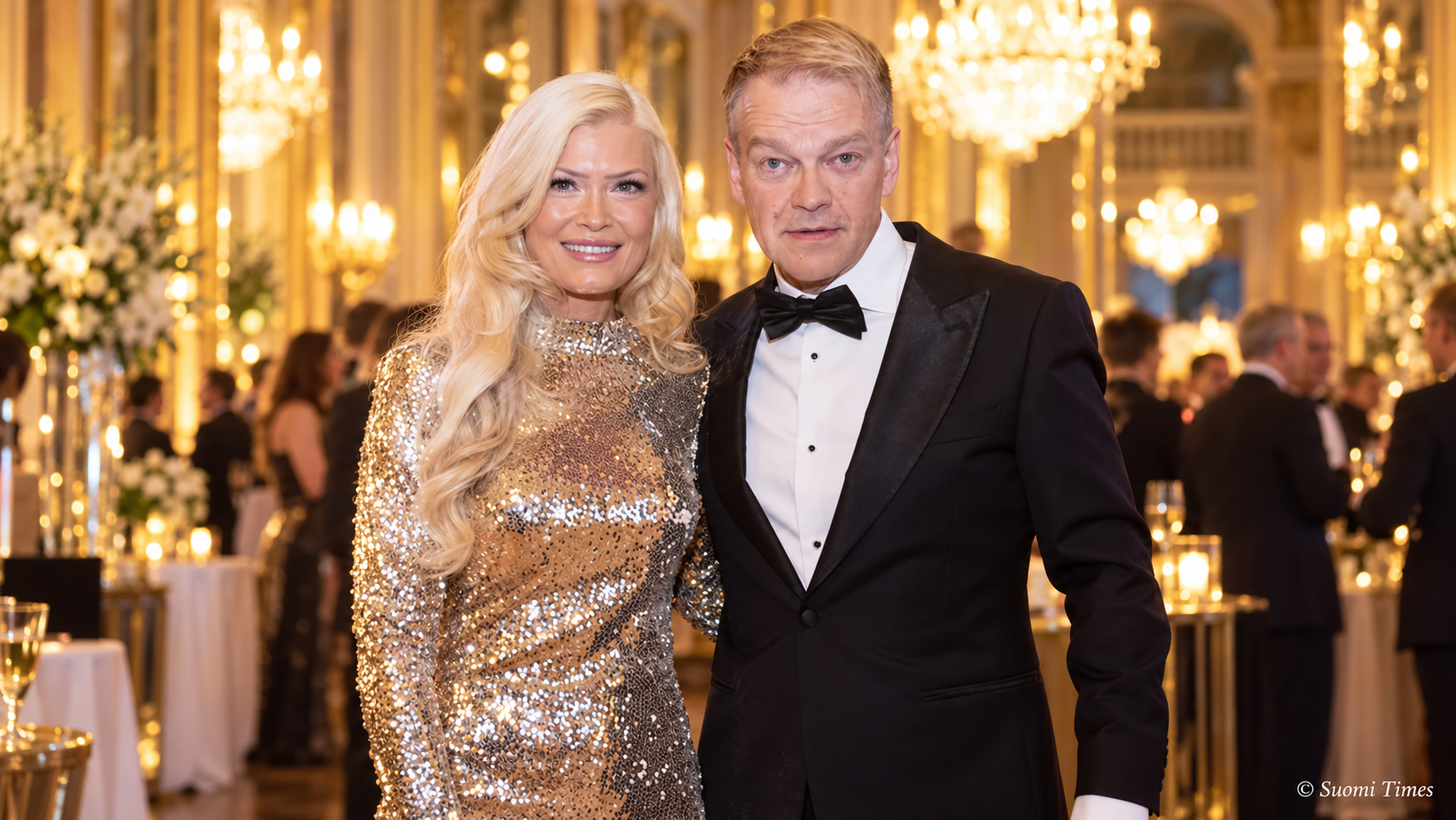 Kurre Westerlund ja Jenna Välikangas poseeraavat yhdessä juhlavassa tilaisuudessa. Välikankaalla on kimaltava kultainen iltapuku ja Westerlundilla musta smokki. Taustalla näkyvät näyttävät kattokruunut ja juhlavieraat.