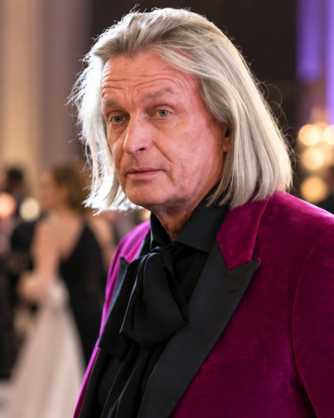 Jukka Rintalan tyylikkäästi viininpunaisessa takissa muotitapahtumassa, taustalla juhlayleisö ja näyttävät asut.