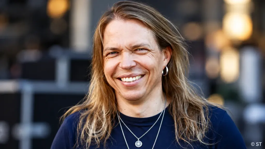 Apocalyptica-yhtyeen sellisti Eicca Toppinen hymyilee kameralle tummansinisessä paidassa, pitkät vaaleanruskeat hiukset olkapäillään. Hänellä on hopeinen korvarengas ja kaulakoru, ja taustalla näkyy pehmeästi sumennettu ympäristö.