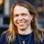 Apocalyptica-yhtyeen sellisti Eicca Toppinen hymyilee kameralle tummansinisessä paidassa, pitkät vaaleanruskeat hiukset olkapäillään. Hänellä on hopeinen korvarengas ja kaulakoru, ja taustalla näkyy pehmeästi sumennettu ympäristö.