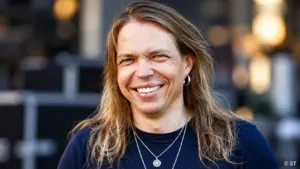 Apocalyptica-yhtyeen sellisti Eicca Toppinen hymyilee kameralle tummansinisessä paidassa, pitkät vaaleanruskeat hiukset olkapäillään. Hänellä on hopeinen korvarengas ja kaulakoru, ja taustalla näkyy pehmeästi sumennettu ympäristö.