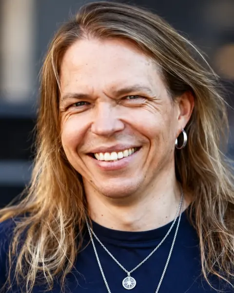 Apocalyptica-yhtyeen sellisti Eicca Toppinen hymyilee kameralle tummansinisessä paidassa, pitkät vaaleanruskeat hiukset olkapäillään. Hänellä on hopeinen korvarengas ja kaulakoru, ja taustalla näkyy pehmeästi sumennettu ympäristö.