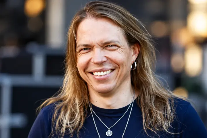 Apocalyptica-yhtyeen sellisti Eicca Toppinen hymyilee kameralle tummansinisessä paidassa, pitkät vaaleanruskeat hiukset olkapäillään. Hänellä on hopeinen korvarengas ja kaulakoru, ja taustalla näkyy pehmeästi sumennettu ympäristö.
