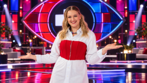 Jenni Poikelus poseeraa Big Brother Suomen uutena juontajana värikkäällä studiolla, taustallaan ohjelman tunnusomainen silmälogo ja näyttävät punasiniset lavavalot.