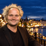 Jani Halme hymyilee Helsingin iltavalaistulla rantakadulla kaupungin valojen loistaessa taustalla.