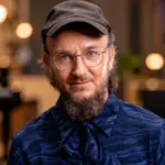 Muusikko Samuli Putro poseeraa tummansinisessä kuviollisessa paidassa, mustassa lippalakissa ja pyöreissä silmälaseissa. Taustalla näkyy pehmeästi sumennettu musiikkistudio, jossa on lämpimiä valoja ja kitara.