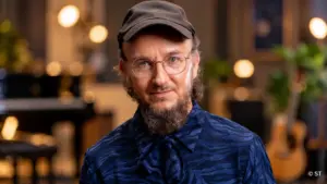 Muusikko Samuli Putro poseeraa tummansinisessä kuviollisessa paidassa, mustassa lippalakissa ja pyöreissä silmälaseissa. Taustalla näkyy pehmeästi sumennettu musiikkistudio, jossa on lämpimiä valoja ja kitara.
