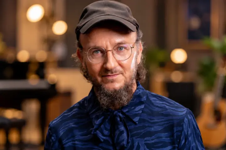 Muusikko Samuli Putro poseeraa tummansinisessä kuviollisessa paidassa, mustassa lippalakissa ja pyöreissä silmälaseissa. Taustalla näkyy pehmeästi sumennettu musiikkistudio, jossa on lämpimiä valoja ja kitara.